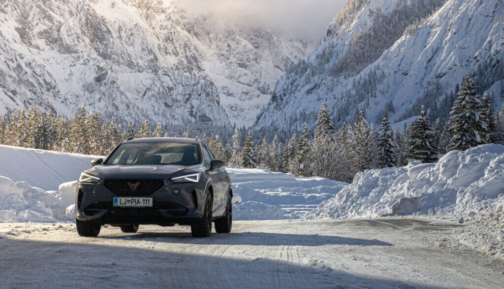 A black SUV with a European license plate is driving on snowy roads in the mountains, surrounded by snowy trees and peaks—a perfect scene showcasing the importance of winter driving and driver safety.